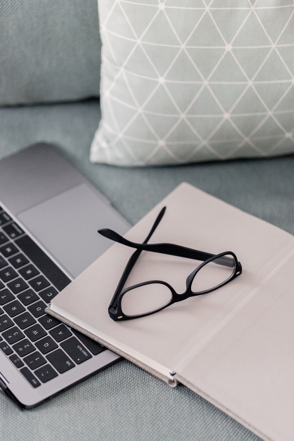 A pair of eyeglasses sitting on top of a notebook and a Macbook laid on a gray couch with a baby blue throw pillow