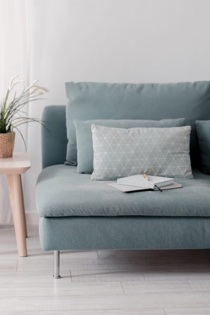 A notebook with a pen on top laid on a baby blue couch with throw pillow and a side table with a plant on it