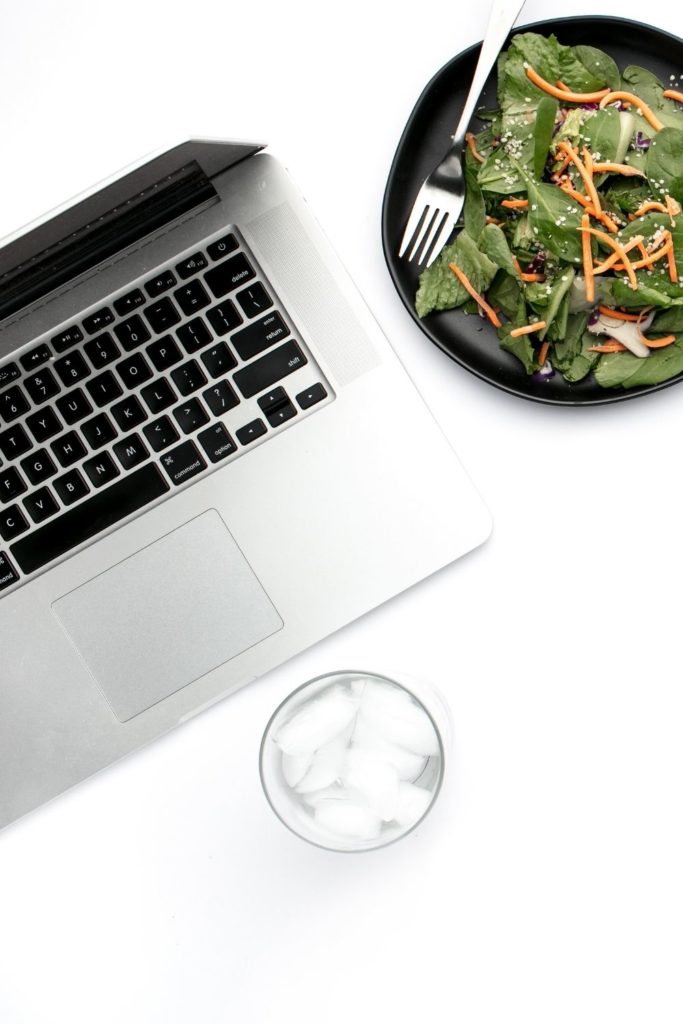 Flat-lay of a Macbook, a glass of iced water and a salad placed side-by-side against a white desk