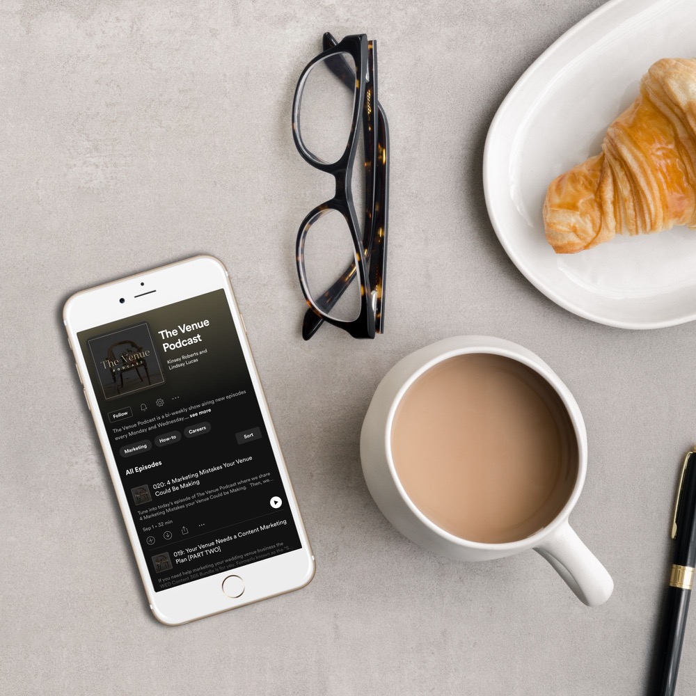 An iPhone showing The Venue Podcast on the screen rests on top of a desk with a pair of glasses, a cup of coffee and a plate of croissant 