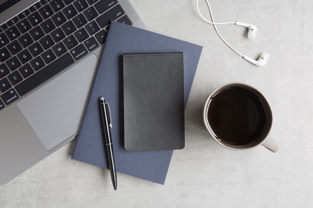 A blue and black notebook with a pen on top laid on a white desk with a Macbook, a cup of coffee and a pair of earphones
