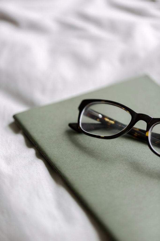 A pair of eyeglasses sitting on top of a thin, green notebook laid on white sheets