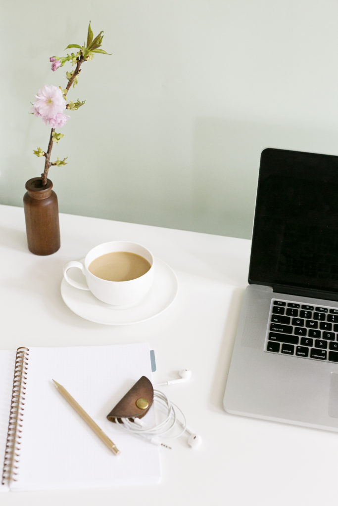 A Macbook laid on a white desk along with a notebook, pencil, a flower on an amber vase and a cup of coffee